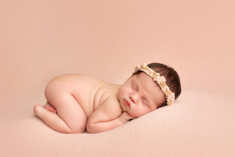 Sleeping newborn curled up on a soft peach backdrop with a floral headband