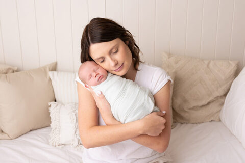 Mum cuddling her swaddled newborn on a white daybed in the studio